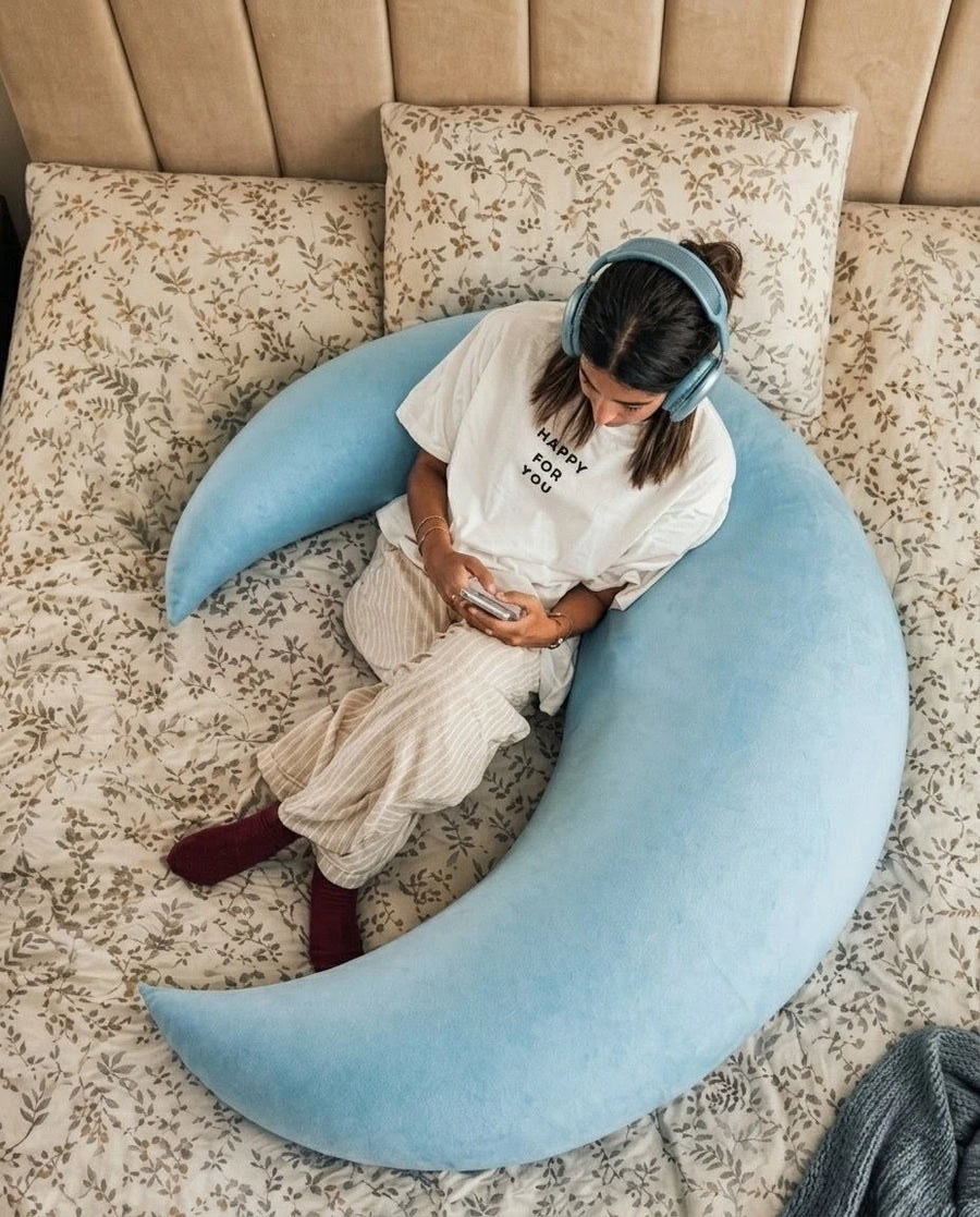 Person sitting on a large blue crescent moon pillow on a bed with floral bedding.