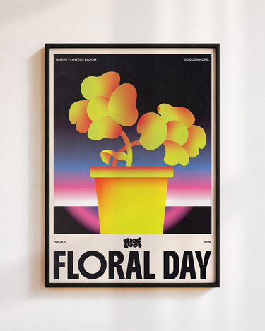 Floral Day poster in a black frame on a white wall: two yellow and orange flowers in a yellow pot against a pink and blue sunset, above the words Floral Day