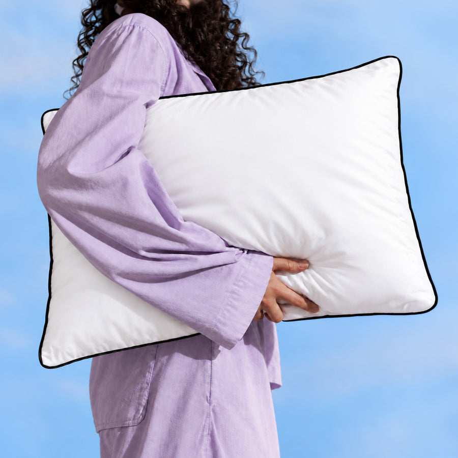 A person in lilac pyjamas carrying a Floof Cloud Pillow against a blue sky